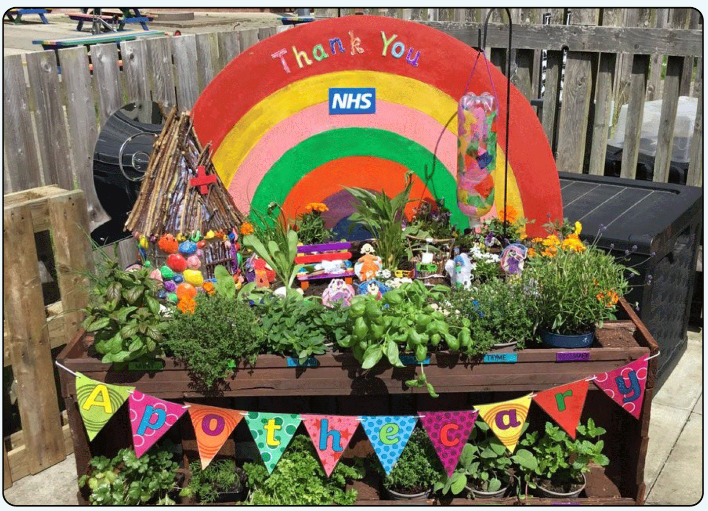 Image of a pocket garden with a series of herbs in pots, a small, thatched roof cottage, park bench made from lollipop sticks, backdrop of painted rainbow with words "thank you NHS" and colourful bunting in foreground with word "apothecary" spelt out.
