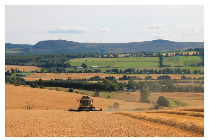 This image shows a large harvester moves through a field of cereal crops under a partly cloudy sky. In the background, the gently rolling hills of the Scottish Lowlands stretch across the horizon, with a patchwork of green fields, tree lines, and scattered farmsteads forming a classic mosaic landscape.