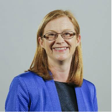 The image shows a photograph of Shirley Anne Somerville, the Cabinet Secretary for Education and Skills.  She is smiling, has shoulder length auburn hair, and is wearing glasses.  She has on a black top and blue suit jacket.