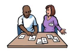 A man and woman are sitting at a table, which has lots of pieces of paper on it. The woman is asking the man to pick one of the pieces of paper and he is pointing at one of them.
