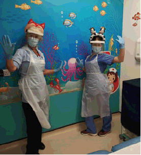 Two female nurses in scrubs and PPE smile and pose in front of an aquarium mural in a hospital