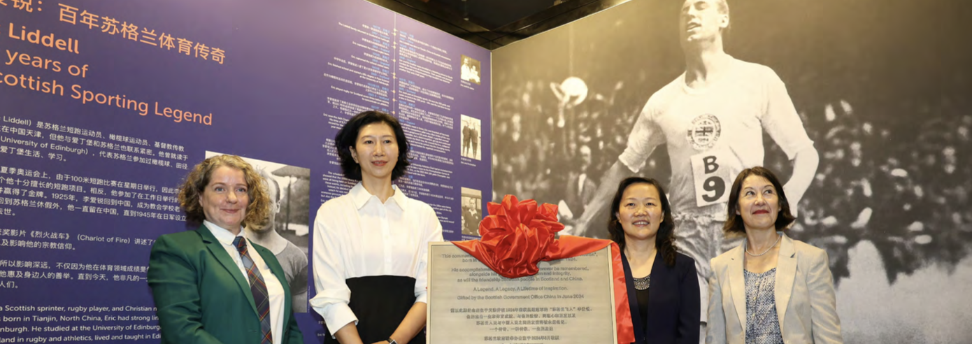 Four women stand by a plaque with a red ribbon in front of an Eric Liddell Exhibition.