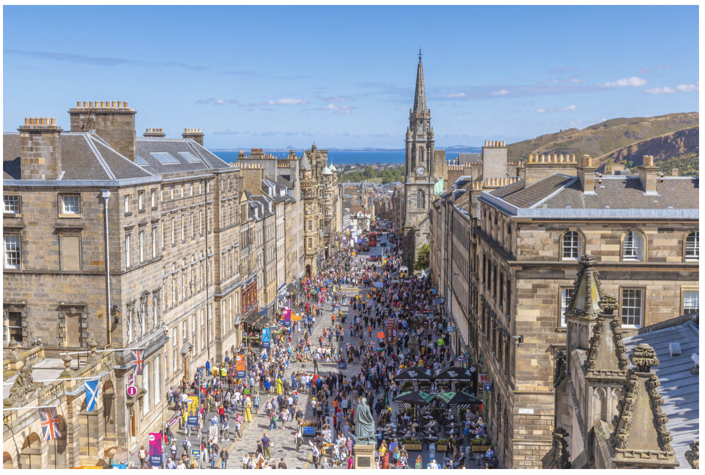 A bustling street scene in Edinburgh with historic stone buildings, festival crowds, colourful flags, and the sea in the distance under a blue sky.