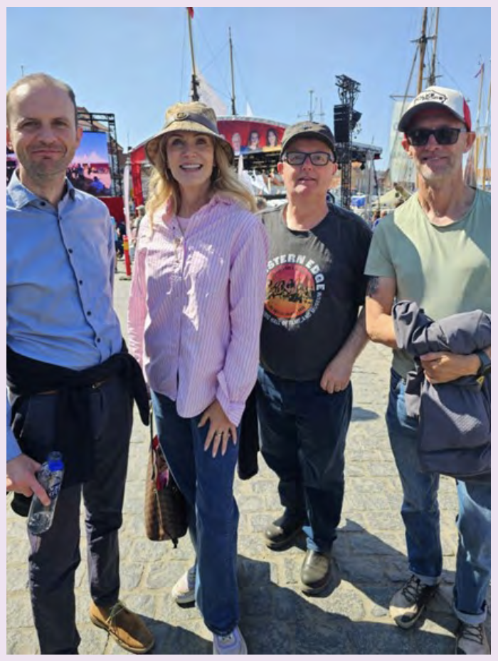 Four people stand smiling at an outdoor event with ships and a stage in the background.