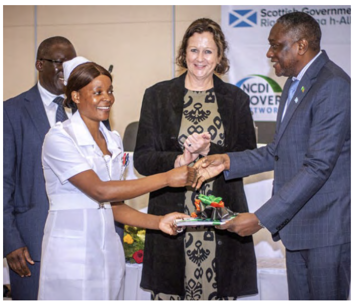 A nurse in white uniform receives an award from a suited man. A woman stands behind them, applauding.