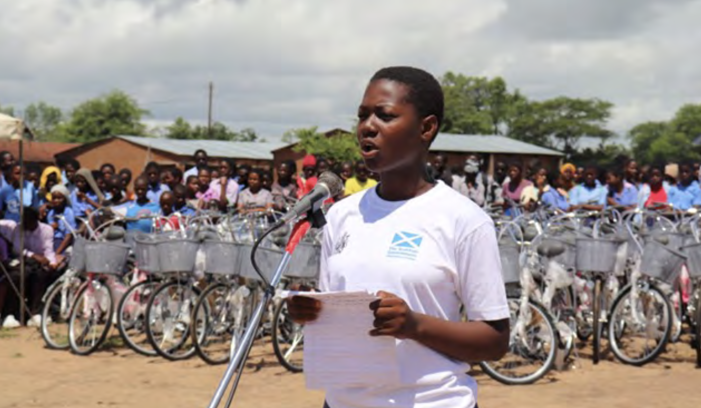 Schoolgirl Alice speaks passionately into a microphone, holding papers.