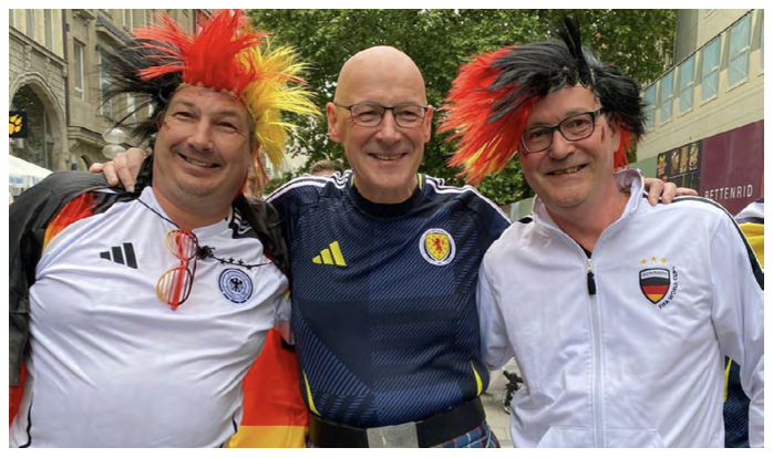First Minister John Swinney wears a Scotland football shirt standing beside two supporters in Germany.