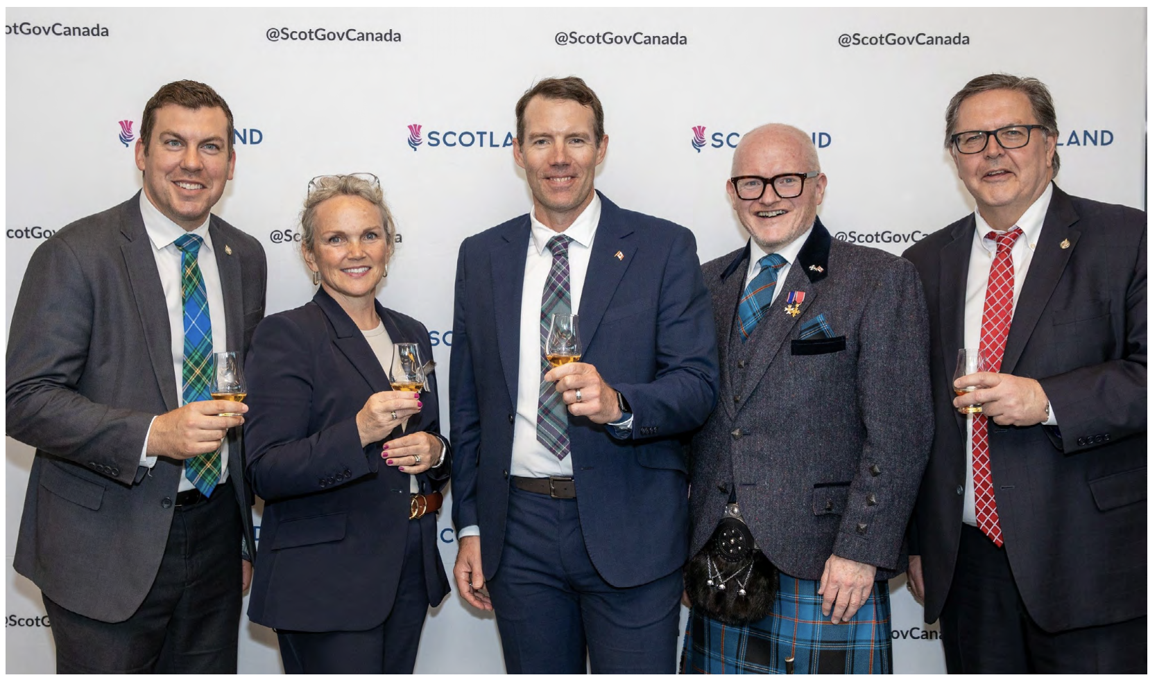 Five people stand in front of a backdrop with Scotland marque, each holding a drink, at a Canadian summer whisky tasting.