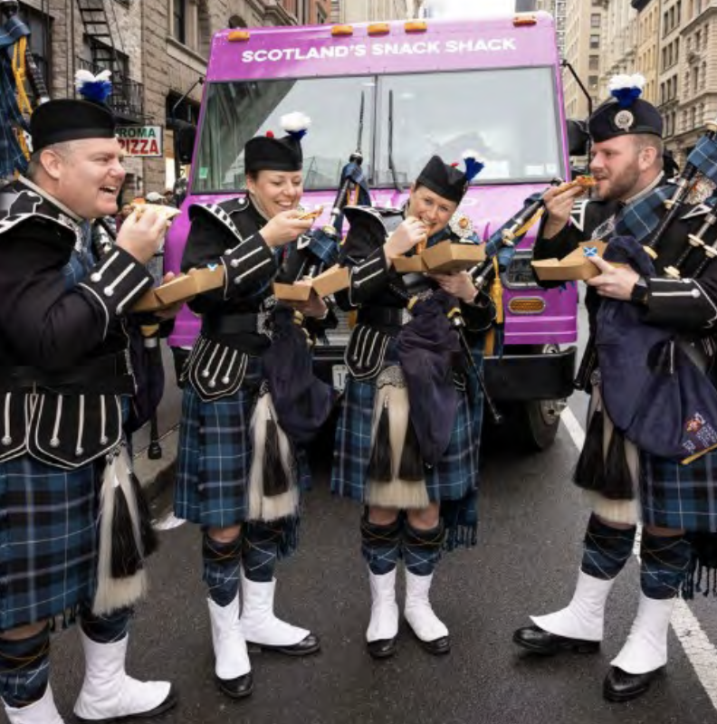 Four performers in traditional Scottish attire enjoy food in front of a truck labelled 'Scotland's Snack Shack' in New York City.