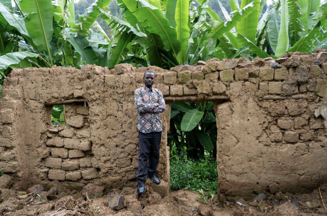 A man stands in the doorway of a crumbling brick wall, showing the damage to his home following floods.