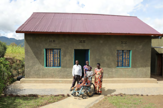 A family of five people, including a young girl in a wheelchair, stand in front of a small house with a red roof.