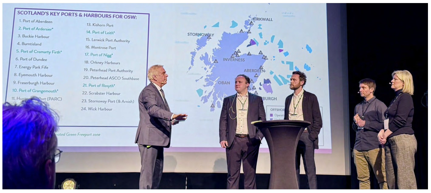 A group of Scottish officials stand on stage in front of a large screen displaying 'Scotland's Key Ports and Harbours' with a detailed map.