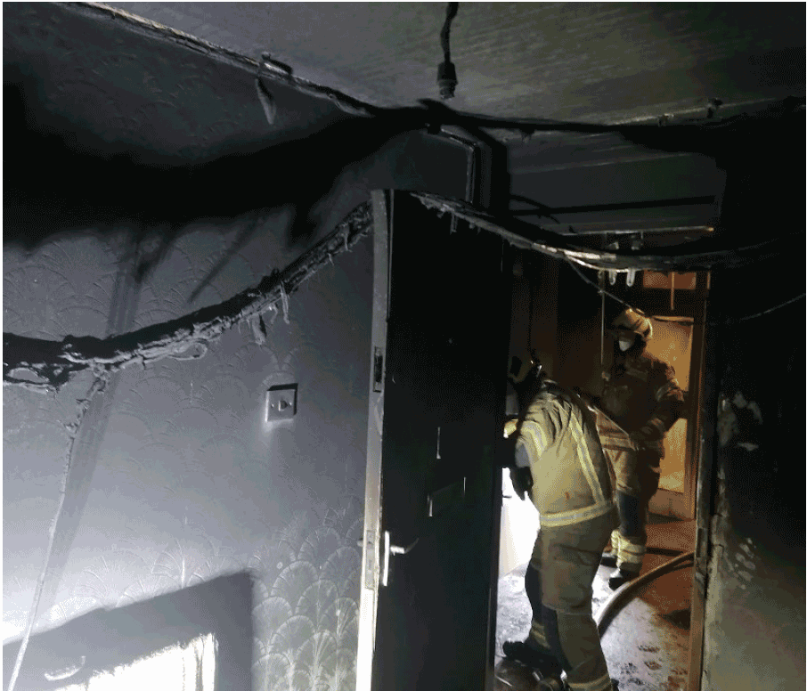 The photograph is of the inside of a room looking toward the entrance door following a fire, a bundle of cables is shown hanging draped across the top of the door having become unattached from the ceiling of the room during the fire.