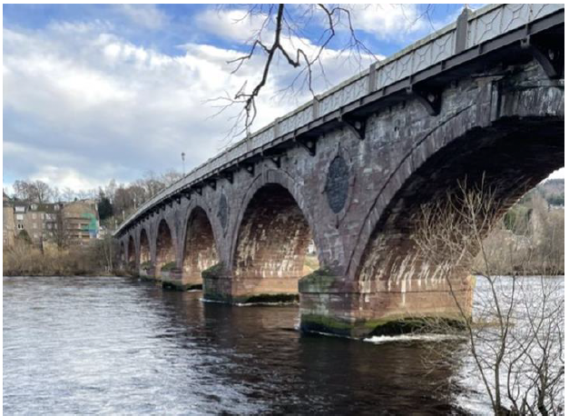 A bridge over the River Tay with trees and buildings in the background
