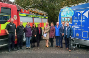 A photo of Cabinet Secretary, Roseanna Cunningham MSP, meeting members of Tillicoultry, Devonside and Coalsnaughton flood action group. The photo has been taken at the community fire station and includes a fire engine and the Scottish Flood Forum’s Flood resilience vehicle in the background. 
