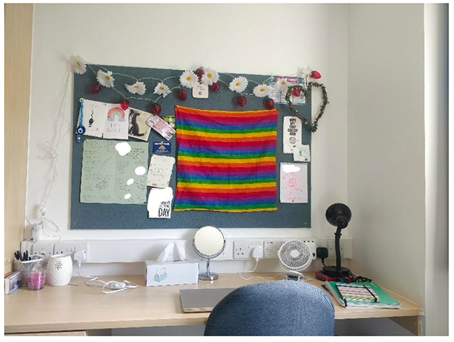 A desk and chair, with a noticeboard above the desk. The noticeboard shows a rainbow scarf, various handwritten notes and a decorative daisy chain.