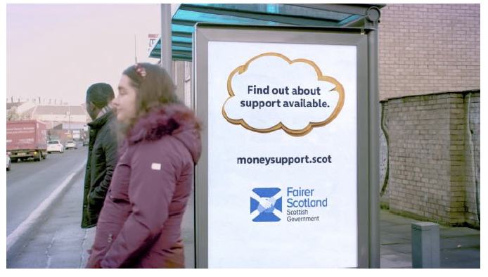 Image shows end frame from the TV advert. A man and woman wait at a bus stop. The billboard on the bus stop reads: Find out about support available. Moneysupport.scot. Fairer Scotland logo.