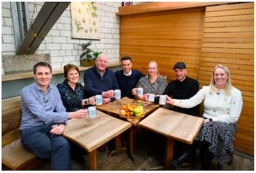 Radio presenter Des Clarke hosting the ‘Be the Early Bird Breakfast Club’. Seven people are sitting around a table in a casual café setting, each holding a ‘Be the Early Bird’ mug and smiling at the camera.