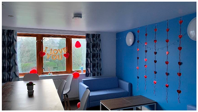 A room with one blue wall and one white wall with windows. Red heart decoration dropping down on the blue wall. A table and chairs, blue couch and coffee table are in view.