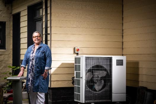 A promotional image from the Heat Pump Heroes Campaign showing an air source heat pump installed in the porch of a house next to a smiling person.